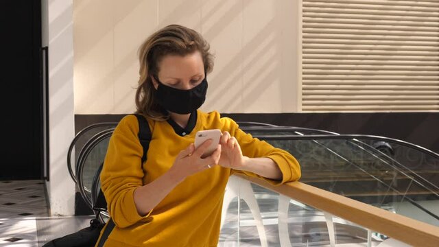 Lonely woman wearing face mask using browsing pn her phone standing in the shopping mall next to escalators