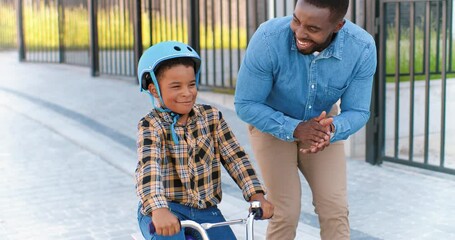 African American handsome father teaching small boy in helmet riding on bike at street in suburb. Little kid riding the bicycle and learning. Schoolboy with daddy. Cheerful son sitting on vehicle.