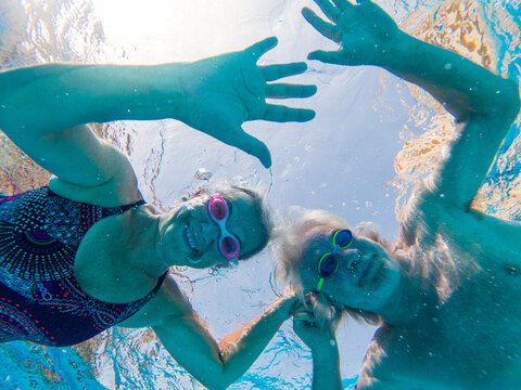 Couple Of Two Happy Seniors Having Fun In The Swimming Pool Swimming And Looking At The Camera Underwater - Mature People Smiling And Enjoying Having A Active Lifestyle