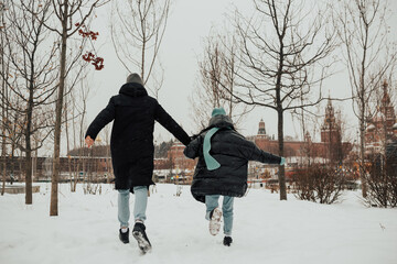 Fototapeta na wymiar Young couple running on a snowy winter field near a big city. Happy and joyful holding hands on romantic date in winter city.
