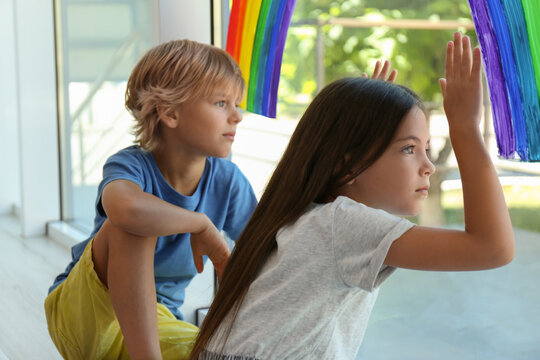 Little children sitting near rainbow painting on window indoors. Stay at home concept
