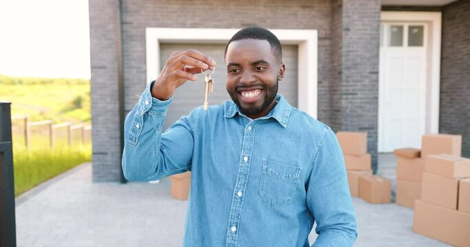 Portrait of cheerful happy African American man smiling to camera and showing key to camera while moving in new home. Outdoor. Male demonstrating keys. Carton boxes on background. Owner of real-estate