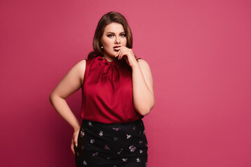 Pensive plus-size model girl with bright makeup and with stylish hairstyle in a red blouse and skirt posing at the pink background, isolated