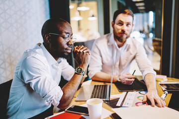 African American boss in optical eyewear reading web news at meeting table with laptop computer, multicultural entrepreneurs working on strategy of partnership using paper documents and netbook