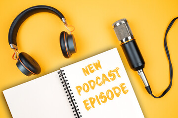 NEW PODCAST EPISODE text on notepad next to headphones and recording microphone, podcasting concept...