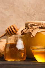 close up view of sweet honey in glass jars with stick on wooden surface near beige concrete wall