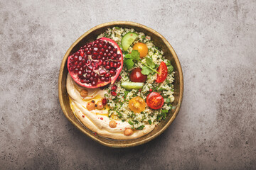 Rustic bowl with couscous salad with vegetables, hummus and fresh cut pomegranate. Middle eastern or Arab style meal with seasonings and fresh cilantro. Healthy Mediterranean dinner, toned image.