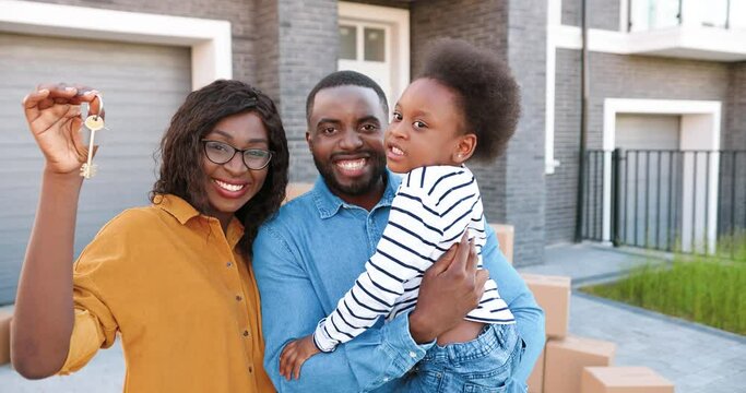 Portrait of happy African American family with small cute girl on hands standing at big house on suburb, smiling and showing key. Joyful mother, father with little pretty daughter. Moving in new home.
