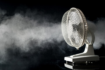Fototapeta Side view of a white electric desktop fan on a black background and reflective floor with visible fog or mist blowing through. Concept of freshness with a fan obraz