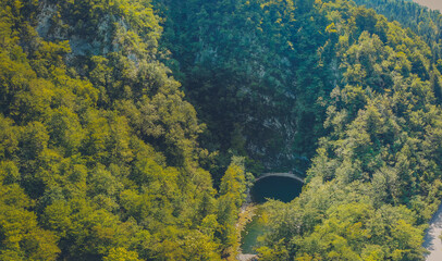 Drone panorama of a round small lake called Divje jezero in Slovenia, surrounded with pebbles and still water on a sunny day with blue skies, hiding between thick green forest.