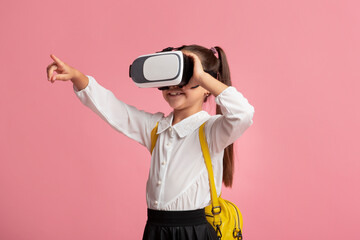 Obraz premium Shocked schoolgirl with virtual reality glasses pointing finger aside on pink background in studio