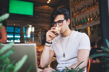 Pensive caucasian male freelancer in eyewear for vision protection drinking coffee while watching video online, serious hipster guy with caffeine beverage browse web page for online course education