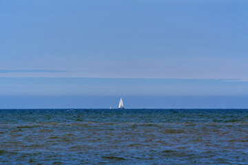 Sailing boat at sea; when zooming into the right half of the horizon one can see vague shapes of ten offshore wind turbines.