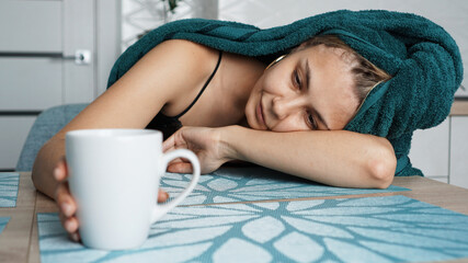 Tired woman sleeping on the table. Beautiful woman in a towel on hair. Hand reaches for the mug....