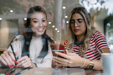Smiling caucasian female using mobile phone for browsing web pages and searching information with friend, positive multiracial girls installing new application on smartphone enjoying 4G in cafe
