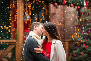 Young man and pretty woman in winter sweaters kissing under the snow in the back yard. Christmas background.