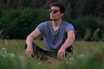 Naklejka na ściany i meble Caucasian Man with Sunglasses Sitting on Meadow and Posing. Young Adult Relaxing in the Nature in Czech Republic.