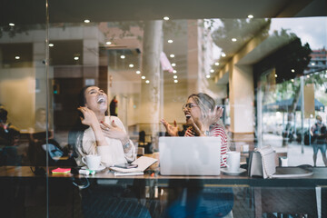 Emotional young female best friends laughing talking to each other on free time break in cafeteria, positive multiracial overjoyed women having fun while discussing plans for online project on leisure