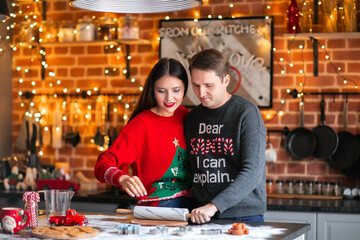 Plakat Young brunette asian woman and white man in Christmas sweaters making cookies together on the kitchen. Lights background.