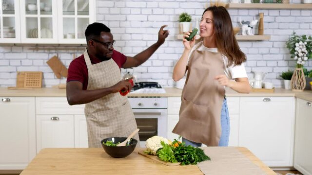 Side view of a mixed race couple enjoying their time together in an apartment, standing in a kitchen, wearing cooking aprons, cooking, dancing, in slow motion. Social distancing and self isolation