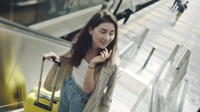 Portrait of young woman going up on escalator with suitcase. Beautiful girl looking tired after train ride, heading to rest. Woman looking around shiny bright station, smiling. Concept of travel.