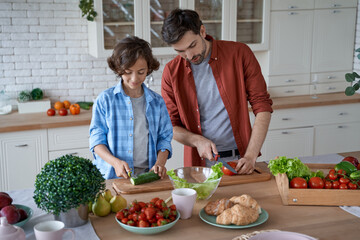 Preparing lunch together. Young father teaching his son how to cut fresh vegetables, preparing salad while standing in the modern kitchen at home
