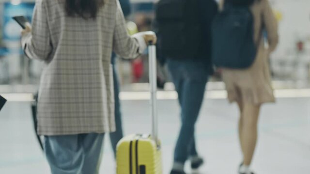 Back view on woman with suitcase walking around railroad station. Girl heading for train tickets for business trip or vacation. Woman moving slowly, checking phone. Concept of travel, transport.