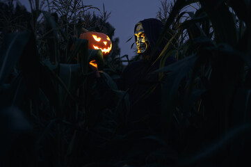 Halloween holiday. A man in a skull mask holds in his hand a glowing Halloween pumpkin in a cornfield at dusk. Faceless. Horror and fear concept.