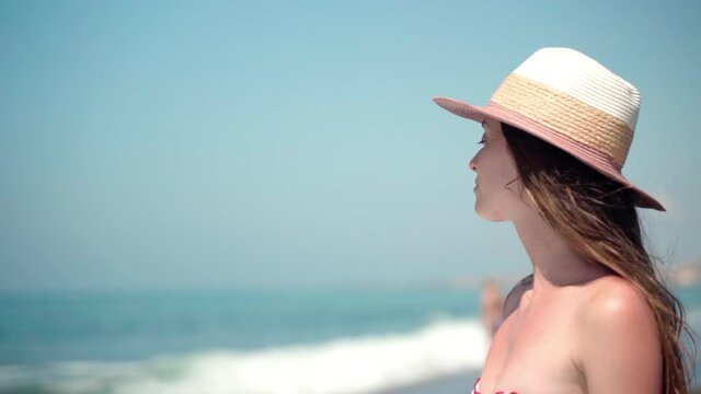 Portrait of concentrated brunette woman in summer hat looking in distance on amazing ocean waves background, sad woman in bikini staying at the beach and relaxing morally. Portrait of pensive female