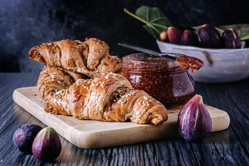 Fresh baked whole grain croissants with a jar of fig homemade marmalade on a wooden cutting board. Front view and dark background.