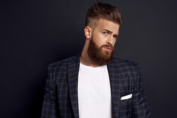 Handsome man with perfect hairdo and beard looking wonderingly at camera while standing against black background