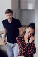 A young scared woman with bruises on her face standing in the kitchen. Drunk man drinking alcohol behind her. The concept of domestic violence and alcoholism