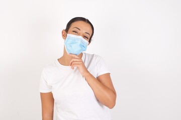 Young arab woman wearing medical mask standing over isolated white background, Optimistic keeps hands partly crossed and hand under chin, looks at camera with pleasure. Happy emotions concept.