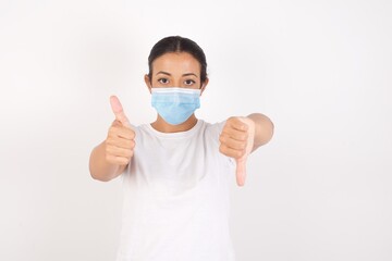 Young arab woman wearing medical mask standing over isolated white background showing thumbs up and thumbs down, difficult choose concept