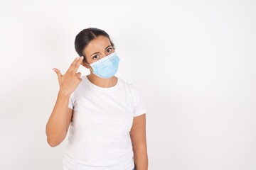 Unhappy Young arab woman wearing medical mask standing over isolated white background makes suicide gesture and imitates gun with hand, curves lips, keeps two fingers on temple