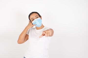 Young arab woman wearing medical mask standing over isolated white background smiling cheerfully and pointing to camera while making a call you later gesture, talking on phone