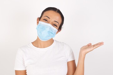 Young arab woman wearing medical mask standing over isolated white background smiling cheerful presenting and pointing with palm of hand looking at the camera.