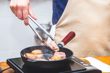 Meat in bacon is fried in a cast-iron frying pan, a man chef in an apron turns the meat over with metal tongs.