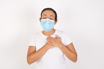 Young arab woman wearing medical mask standing over isolated white background smiling with hands on chest with closed eyes and grateful gesture on face. Health concept.