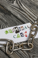 The phrase Criminal case made of letters cut from a magazine and pasted on a sheet of paper. There is a metal chain nearby. On brushed pine boards painted black and white.