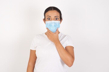 Young arab woman wearing medical mask standing over isolated white background Looking fascinated with disbelief, surprise and amazed expression with hands on chin