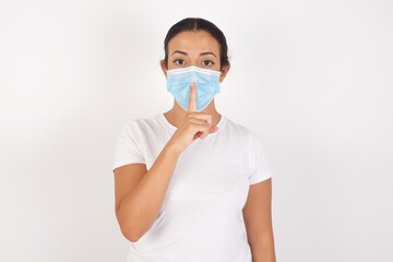 Young arab woman wearing medical mask standing over isolated white background asking to be quiet with finger on lips. Silence and secret concept.