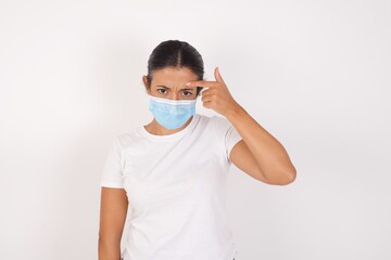 Young arab woman wearing medical mask standing over isolated white background pointing unhappy at pimple on forehead, blackhead  infection. Skincare concept.