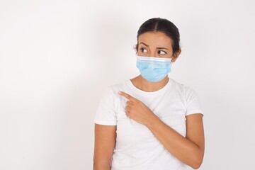 Young arab woman wearing medical mask standing over isolated white background. Pointing aside worried and nervous with forefinger, concern and surprise concept.
