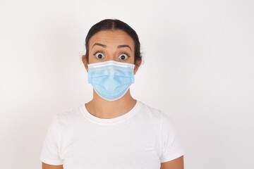 Oh my God. Surprised  Young arab woman wearing medical mask standing over isolated white background stares at camera with shocked expression exclaims with unexpectedness,