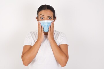 Studio shot of scared terrified Young arab woman wearing medical mask standing over isolated white wall shocked with prices at shop, being short of money to buy something, People and human emotions 