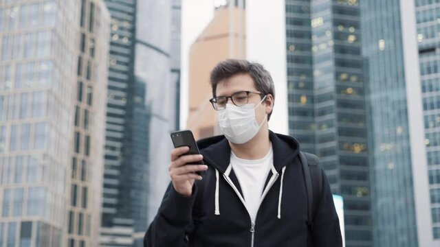 Man wearing a mask walking with a phone on background of skyscrapers in business centre. Man dressed in casual clothes holding a phone walking 