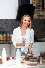 Young woman standing in the kitchen at home and cooking with enjoyment bakery products of flour, milk, cocoa, sugar and eggs. Funny portrait of happy girl chef