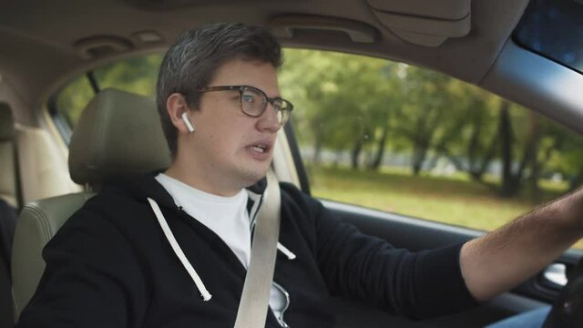 Man in eyeglasses using earpods in a car, dressed in casual clothes. Gimbal shot of man receiving a call through earpods, sitting in the car on background of green city trees