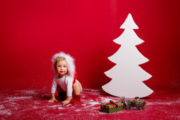 A little girl in a red dwarf hat sits on the floor near the decoration of a white Christmas tree